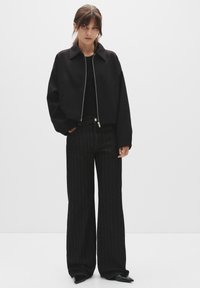 Jachetă neagră scurtă cu guler înalt și fermoar argintiu, combinată cu pantaloni negri cu dungi verticale, cu talie înaltă. Material neted și design ajustat.