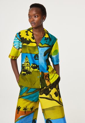 Vrouw met kort haar, gekleed in een kleurrijk shirt met een abstract patroon en korte mouwen, en bijpassende losse broek, staand met haar handen in de zakken.