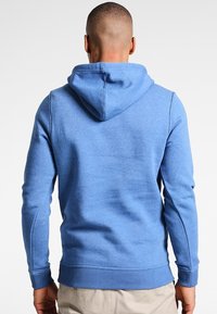 Ljusblå hoodie av mjukt tyg, med dragsko i huvan, ribbade ärmar och en kenguruficka med platta sömmar.