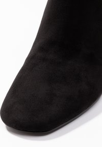 Primer plano de la puntera de un zapato de ante negro con forma de cuadrado redondeado sobre un fondo blanco.