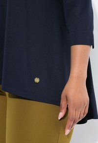 Tummansininen tunika, jossa on lyhyet hihat ja kultainen varuste alareunassa. Yhdistetty oliivinvihreisiin leggingsseihin. Sujuva rakenne.