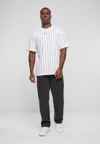 Mand står iført en hvid stribet t-shirt med rødt logo, sorte bukser og hvide sneakers foran en ensfarvet lys baggrund.