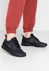Personne portant des baskets Nike Air Max noires et un pantalon de jogging rouge debout sur un sol blanc lisse.