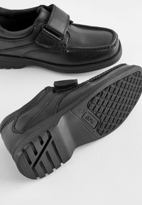 Chaussures en cuir noir avec une sangle Velcro, bout arrondi et semelle en caoutchouc texturée présentant un motif en losanges pour une meilleure adhérence.