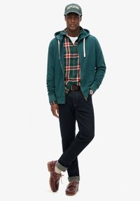 Sudadera con cremallera color teal sobre una camisa de cuadros en rojo y verde, jeans oscuros, zapatos marrones con un puño crema, y una gorra de béisbol verde con texto blanco.