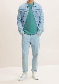 Homme portant une veste en denim bleu clair, un pull texturé vert sarcelle, un jean bleu clair retroussé, des chaussettes blanches et des baskets blanches, les mains dans les poches.