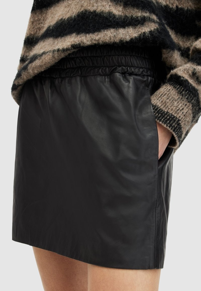 Femme portant une mini-jupe en similicuir noire avec une ceinture élastique et la main dans la poche, associée à un pull duveteux rayé brun et noir.