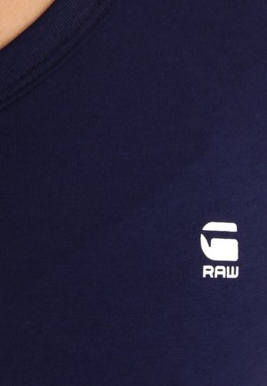Close-up of navy blue fabric with white stylized "G RAW" logo printed on it.