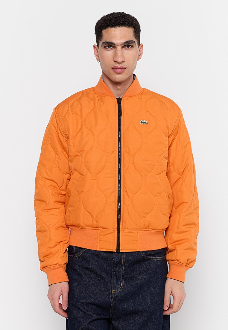 Veste bomber orange matelassée avec fermeture éclair à l'avant, poignets et ourlet côtelés. Présente un petit logo vert sur la poitrine et des poches latérales.