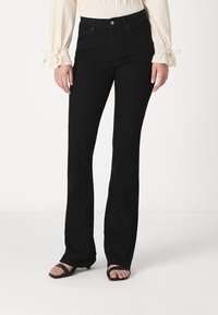 Femme portant un jean noir bootcut, un chemisier crème à manches longues avec des détails en forme de nœud aux poignets, et des sandales noires à lanières, se tenant devant un fond blanc.