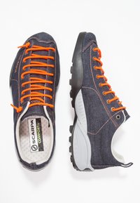 Tummansinisiä farkkukengät, joissa oranssit nauhat. Kumipohja, jossa pito-kuvioita, pehmustettu sisäosa ja harmaat yksityiskohdat pohjassa.