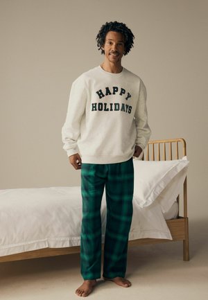Hellgrauer Sweatshirt mit "FROHE FEIERTAGE" in dunkler Schrift, kombiniert mit grünen Karohosen, barfuß neben einem Bett stehend.