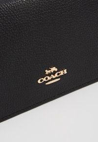 Portefeuille noir en cuir avec une surface texturée. Il arbore un logo doré "COACH" avec un motif de calèche. Conception simple et élégante.