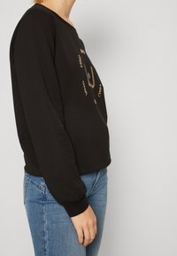 Czarna bluza z długimi rękawami i okrągłym dekoltem, zdobiona złotym nadrukiem graficznym. W zestawieniu z jasnoniebieskimi dżinsami, podkreślającymi dopasowanie i styl.