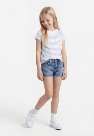 Mladé dievča sa usmieva dolu, má na sebe biele tričko s pruhmi, rifľové šortky, biele ponožky a biele tenisky, stojí na jednej nohe.