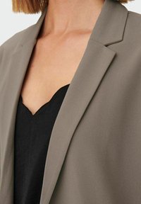 Grijze blazer met een gladde textuur en inkepingen op de revers,laagjes over een zwarte top met een geschulpte halslijn. Close-up van de stof en het ontwerp.