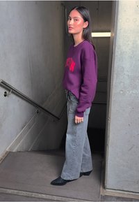 Fioletowa sweatshirt z napisem "LOGO" w czerwonym kolorze, szare dzwonowe dżinsy i spiczaste czarne botki, w tle schody betonowe.