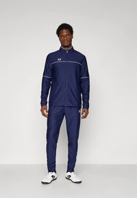 Temno modra atletska trenirka iz gladke tkanine, z zadrgo na jakni in belimi poudarki ter ujemajočimi hlačami. Oblečena s belimi športnimi copati.