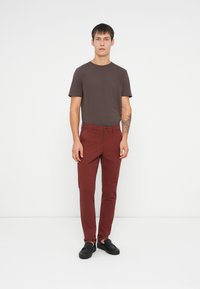 Smeđi majica kratkih rukava u kombinaciji s maroon uskim hlačama. Model nosi crne cipele na vezanje, stoji na neutralnoj pozadini.