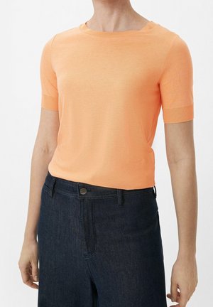 Vrouw draagt een perzikkleurige korte mouwen gebreide top, ingestopt in een donkerblauwe high-waisted spijkerbroek met knoopsluiting.