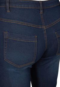 Temno modri jeans iz denima z oprijeto zasnovo, ravno krojenimi hlačnicami in oranžnim šivanjem na žepih ter šivih. Zadnji pogled z dvema žepoma.