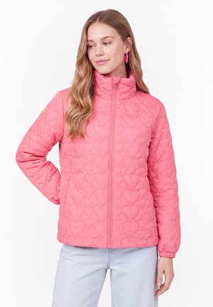 Jeune femme portant une veste matelassée rose avec des motifs de cœurs, debout avec une main dans la poche et regardant sur le côté.
