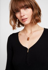 Zwarte geribbele top met een diepe V-hals en een rits aan de voorkant. Bevat een delicate gouden ketting met een rechthoekige hanger.