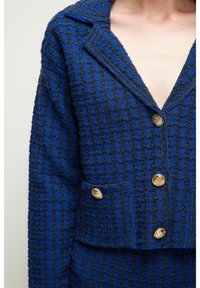 Blazer texturizado azul con un patrón de pata de gallo, cuello con muesca y botones dorados. El material parece tejido con un diseño estructurado.