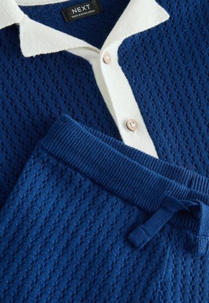 Modra pletena jopica z belim ovratnikom, z zaponko z gumbi. Udobna teksturirana tkanina z vzorčastim dizajnom, združena z ujemajočimi modrimi trenirkami.