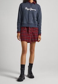 Felpa blu scuro con testo bianco, gonna a camicia in tartan rosso e stivali neri con lacci. L'outfit combina elementi casual e a strati.