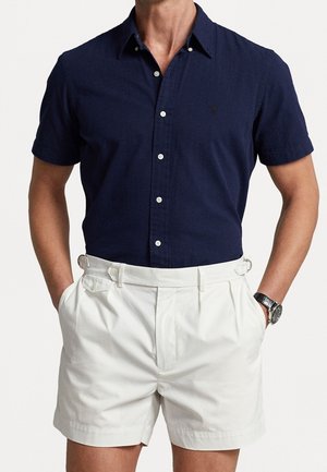 Hombre que lleva una camisa azul marino de manga corta abotonada con botones blancos y pantalones cortos blancos plisados con ajustadores laterales y bolsillos.