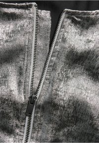 Metallisch silberner Stoff mit einer glatten Textur, der in der Mitte einen schwarzen Reißverschluss aufweist. Der Reißverschluss befindet sich vor einem kontrastierenden dunklen Hintergrund.
