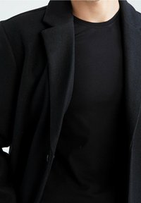 Schwarze Außenjacke aus weichem Stoff mit Reverskragen und taillierter Passform. Darüber wird ein schwarzes, glatt strukturiertes Hemd getragen.