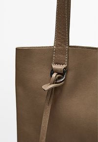 Borsa tote in pelle marrone chiaro con una superficie liscia, dotata di una lunga tracolla attaccata con un anello metallico nero e dettagli di cucitura visibili.
