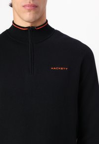 Sudadera negra con cremallera y cuello alto, con una fina franja naranja y un logo naranja de "HACKETT" en el pecho. Textura de punto suave.