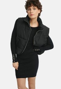 Czarna kamizelka puchowa z zapięciem na zamek, z wysokim kołnierzem i przednimi kieszeniami, noszona na czarnej sukience z prążkowanej tkaniny z akcentami złotych guzików.