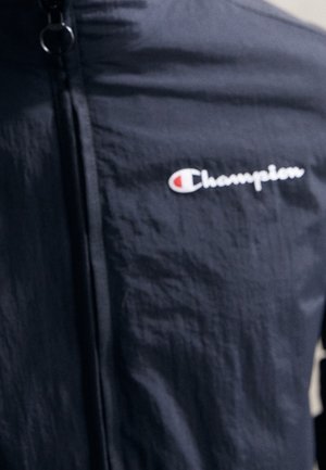 Schwarze Zip-up-Jacke mit dem "Champion"-Logo, das in Weiß und Rot auf der linken Brust gestickt ist.