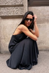 Mustaton hihaton mekko, jossa on virtaava, teksturoitu hame. Mustat aurinkolasit ja sileä hiustekstuuri täydentävät asua. Kaupunkimainen betonitausta.