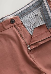 Pantaloni de culoare ruginie, cu o textură fină, având un fermoar și închidere cu buton, precum și o căptușeală interioară cu dungi.