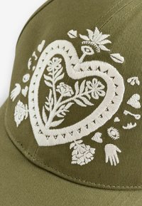 Oliveiroheline nokamüts, millel on valge tikkimine südamekujulise mustri, lillemotiivide, käte ja dekoratiivsete elementidega.