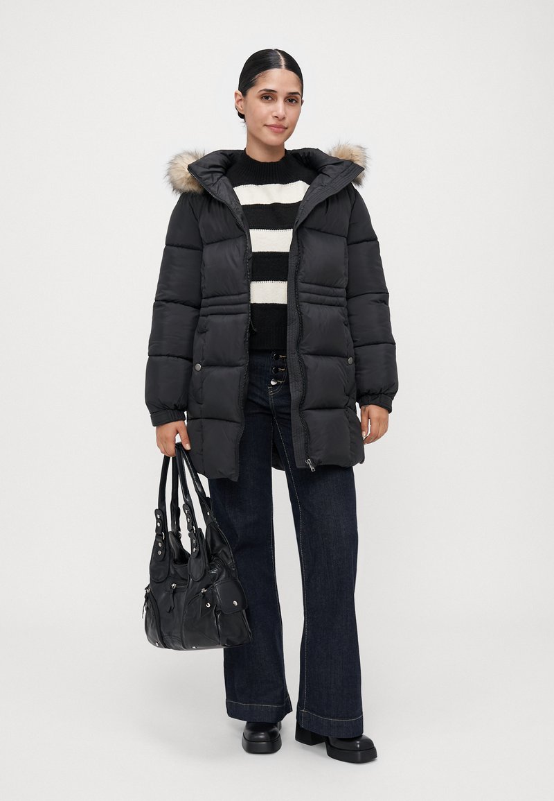 Mujer con un abrigo de plumas negro con capucha de piel, suéter a rayas negro y blanco, jeans oscuros de pierna ancha, zapatos negros y sosteniendo un bolso negro.