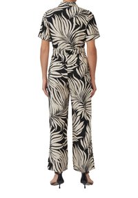 Zwart en beige jumpsuit met een bladpatroon, korte mouwen, een getailleerde taille, wijde benen en een soepele stoftextuur. Zwarte hakken zijn zichtbaar.