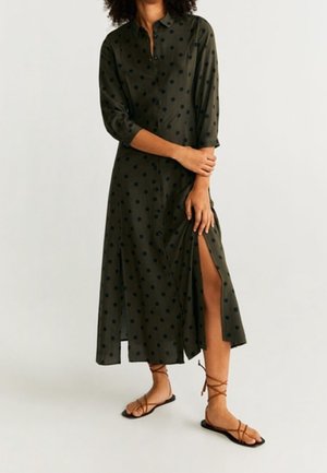 Robe chemise longue vert olive à pois noirs, boutonnée sur le devant, manches trois-quarts, fente latérale ; associée à des sandales marron à brides.