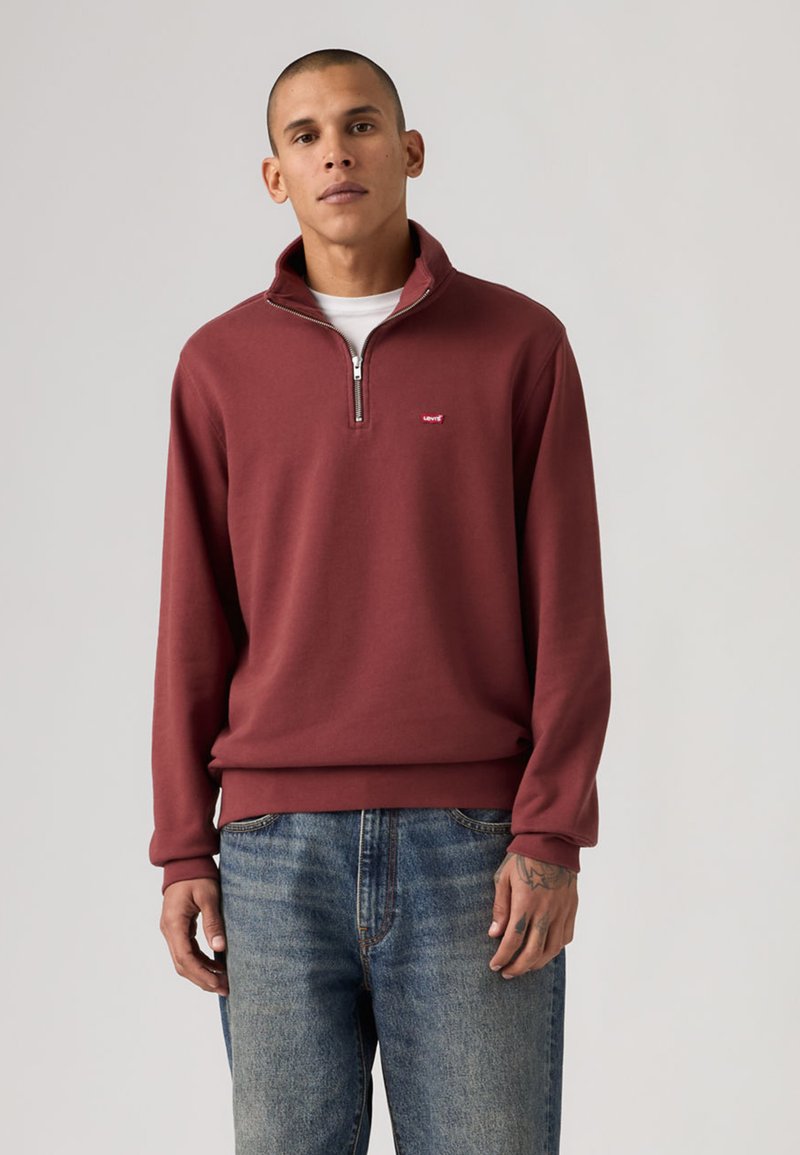 Άνδρας που φοράει μπορντό πουλόβερ με φερμουάρ μέχρι τη μέση πάνω από λευκό πουκάμισο και μπλε τζιν, στέκεται μπροστά σε απλό φόντο.