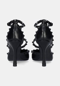 Sandalias de tacón alto negras con punta afilada, acentos de tachuelas y correas en el tobillo. Presentan un acabado brillante y tacones de aguja estilizados.