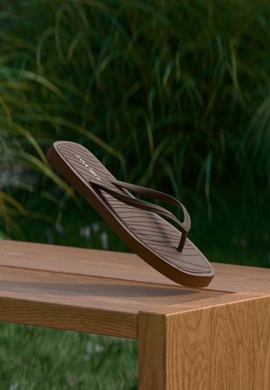 Barna flip-flop szandál egyensúlyozva egy fa asztal szélén, a háttérben magas zöld fűvel, szabadtéren.