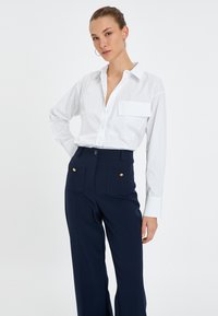 Camisa blanca de botonadura con un bolsillo en el pecho, combinada con unos pantalones de talle alto azul marino que presentan acentos de botones dorados y grandes bolsillos frontales.
