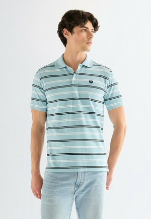 Giovane uomo che indossa una polo a righe azzurro chiaro e jeans azzurri, guarda verso destra su uno sfondo bianco uniforme.