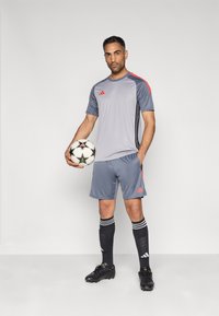 Γκρι αθλητική φανέλα και σορτς με κόκκινες λεπτομέρειες, συνδυασμένα με μαύρες κάλτσες με λευκές ρίγες και μαύρα παπούτσια ποδοσφαίρου, κρατώντας μια μπάλα ποδοσφαίρου.