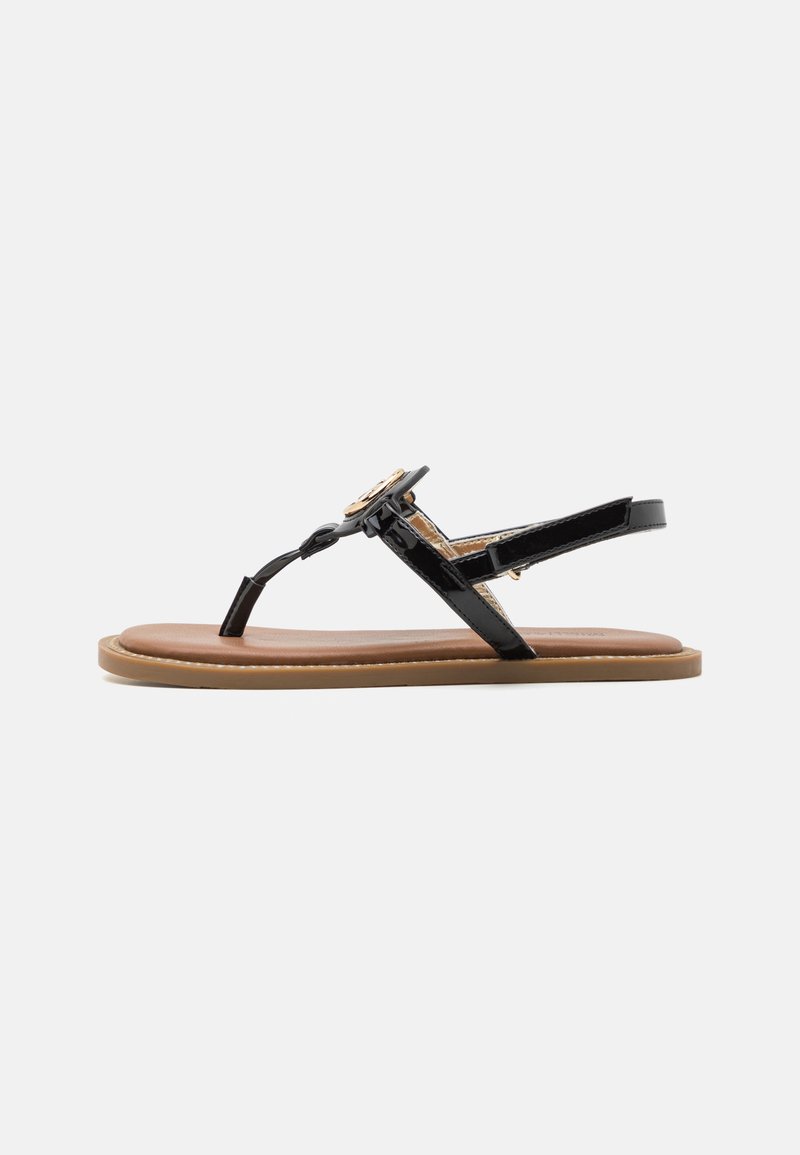 Svart lackläder sandal med tongsstil, prydd med en cirkulär gyllene detaljer och en beige vadderad innersula, placerad på en ljusbrun sula.
